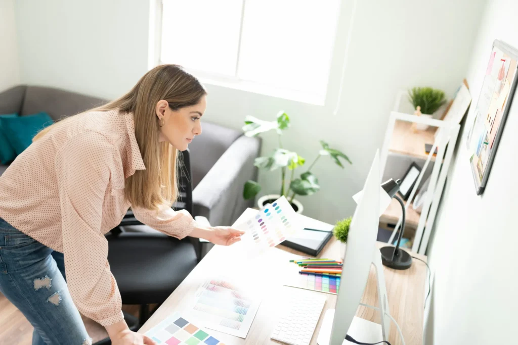 Designer in light pink blouse examining color swatches while leaning over desk with computer monitor. Workspace features color charts, colorful pencils, and potted plants in bright, naturally lit home office with gray sofa.