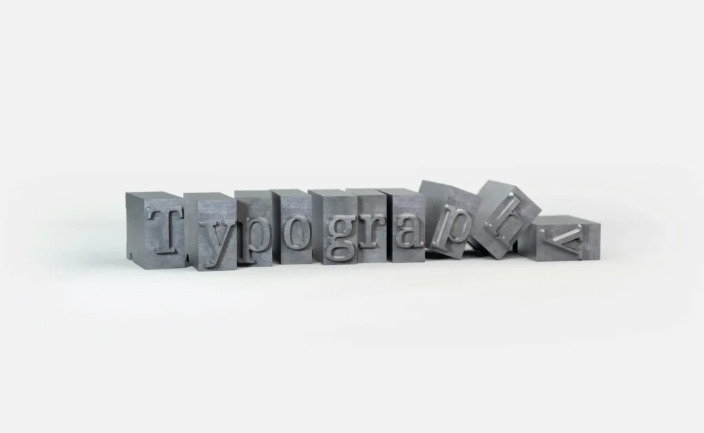 Metal letterpress printing blocks spelling 'Typography' on a white background, with the last few blocks toppling over. The blocks have a brushed silver appearance and cast subtle shadows.