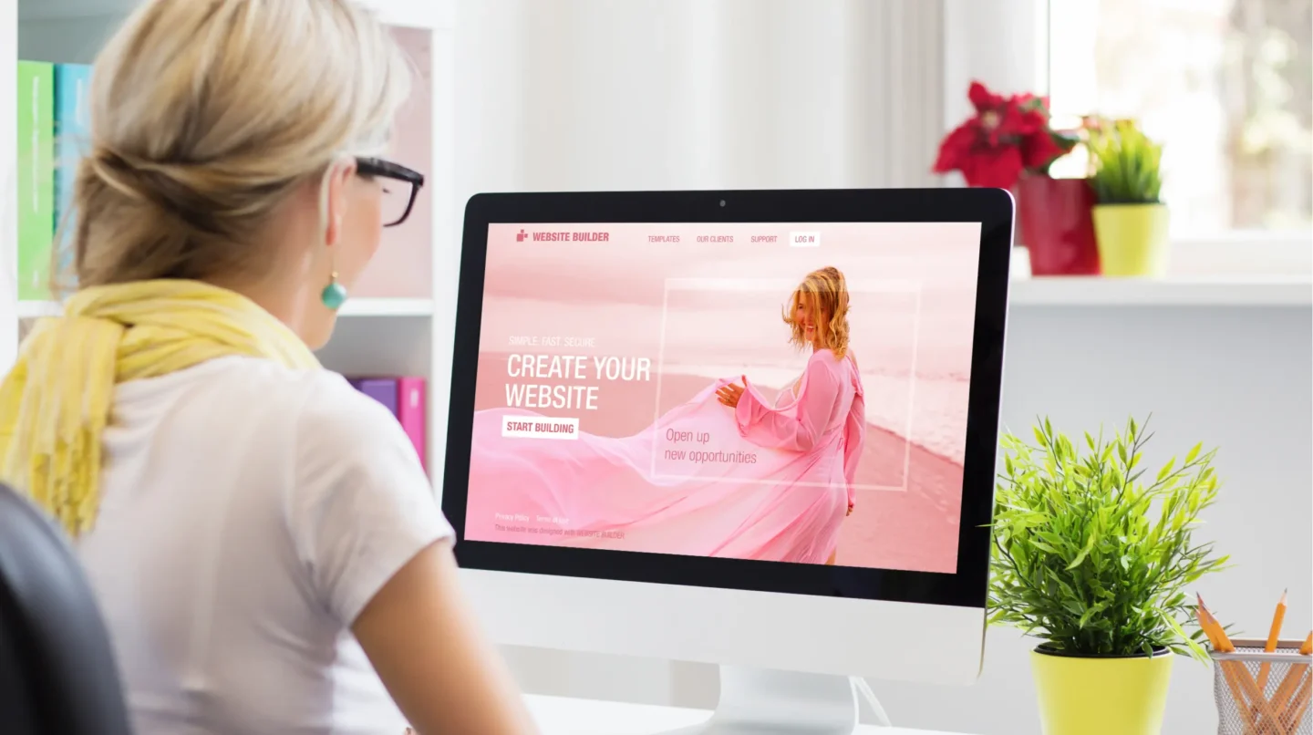 Alt Description: Woman viewing website builder homepage on desktop computer displaying Create Your Website call-to-action in bright home office.