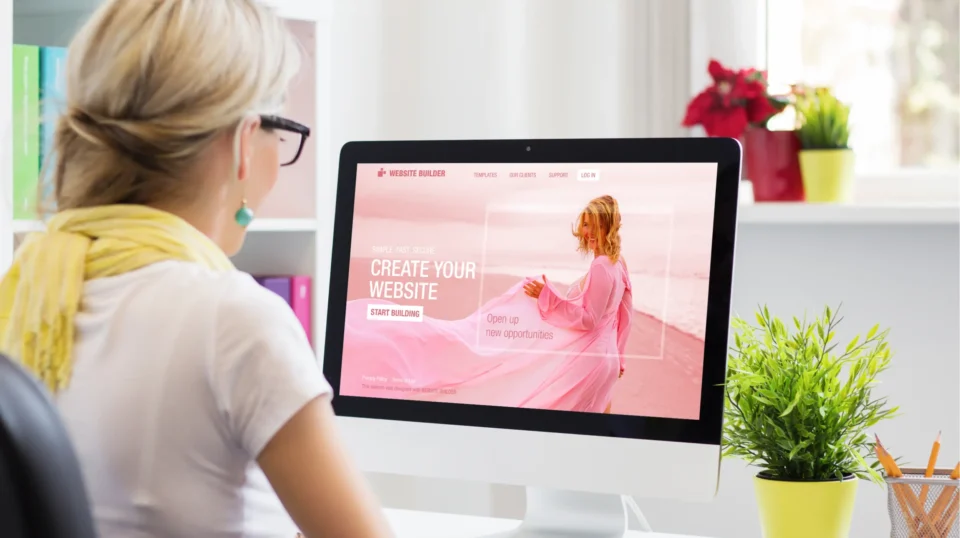 Alt Description: Woman viewing website builder homepage on desktop computer displaying Create Your Website call-to-action in bright home office.