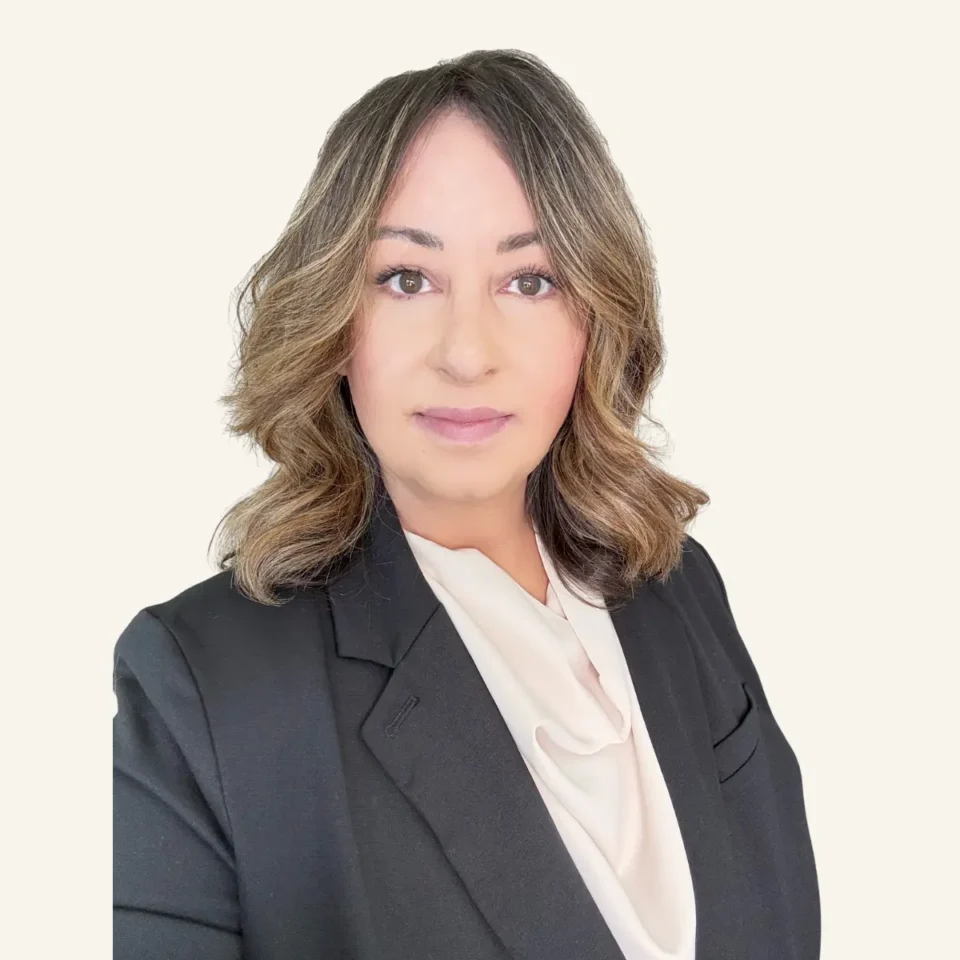 Professional headshot of Erika Huber, business owner, with wavy brown hair wearing black blazer and white blouse against neutral background.