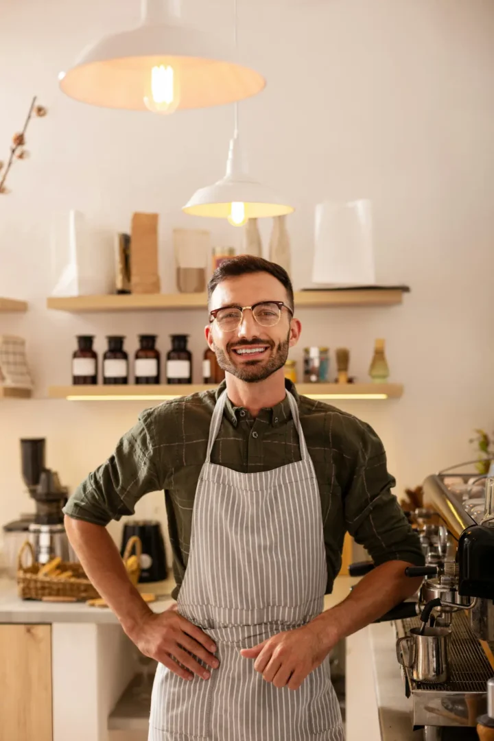 Small business owner in striped apron standing behind coffee shop counter with espresso machine and pendant lighting.