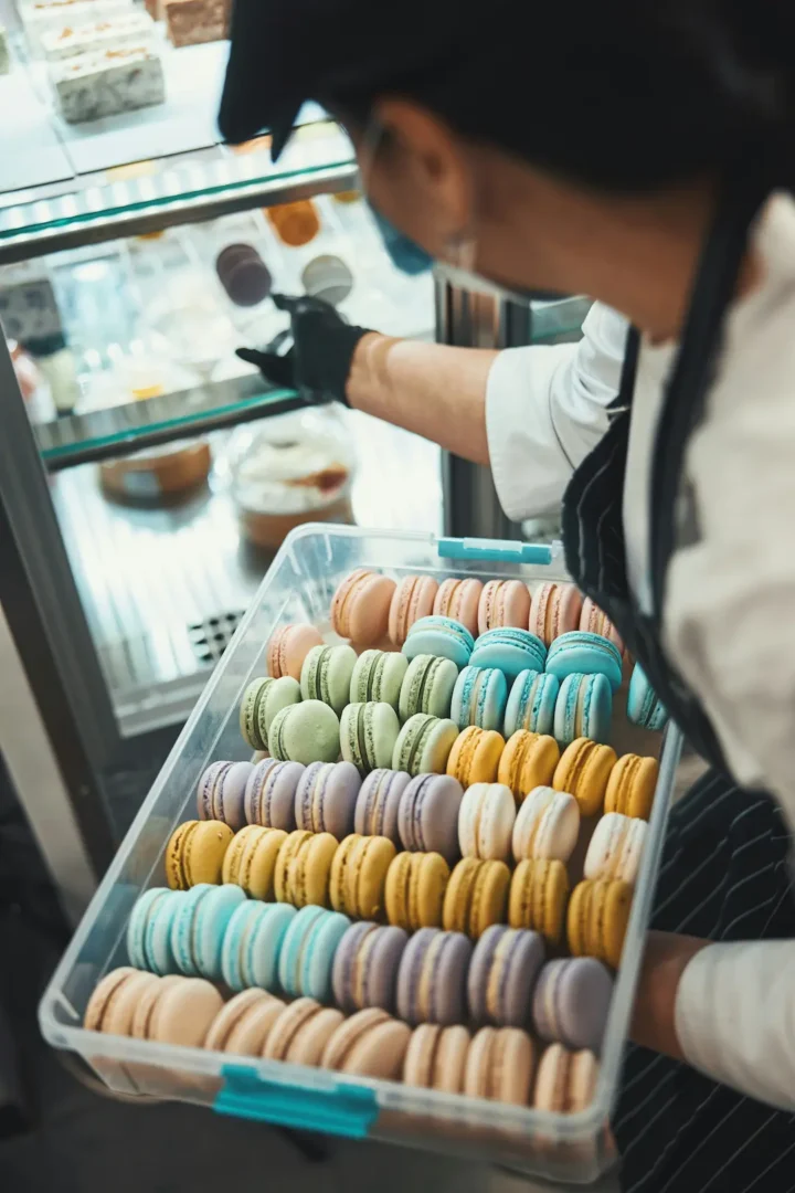Baker in apron holding tray of colorful macarons in pastel shades at bakery display case.