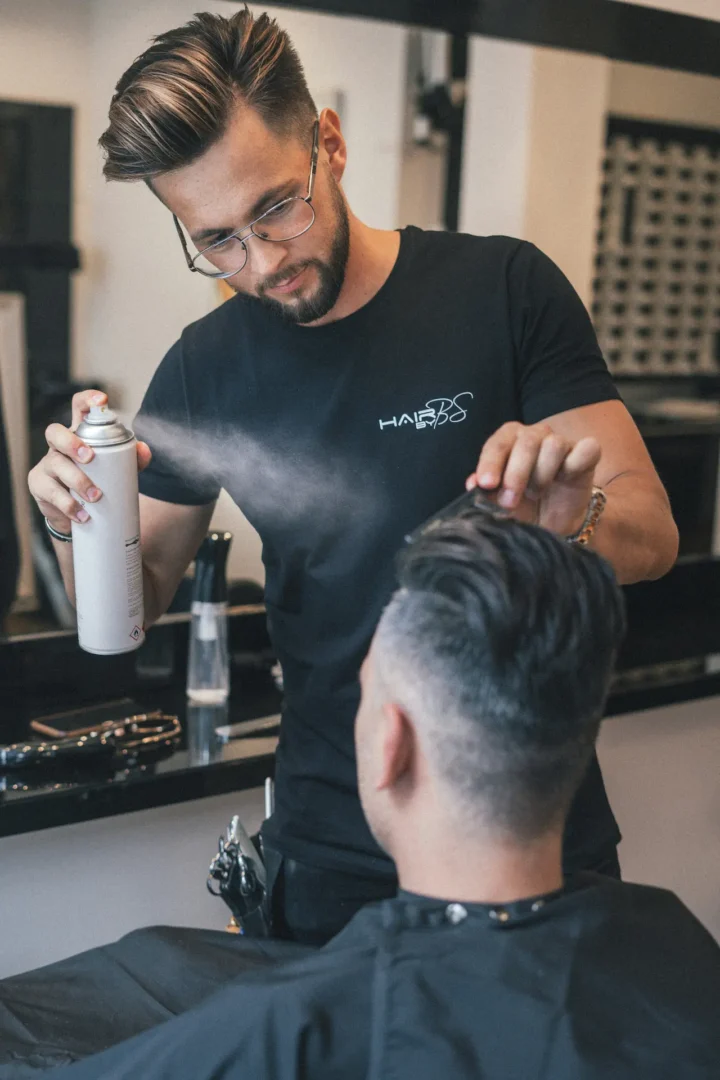 Barber wearing glasses applying hairspray while styling client's hair in modern salon setting.