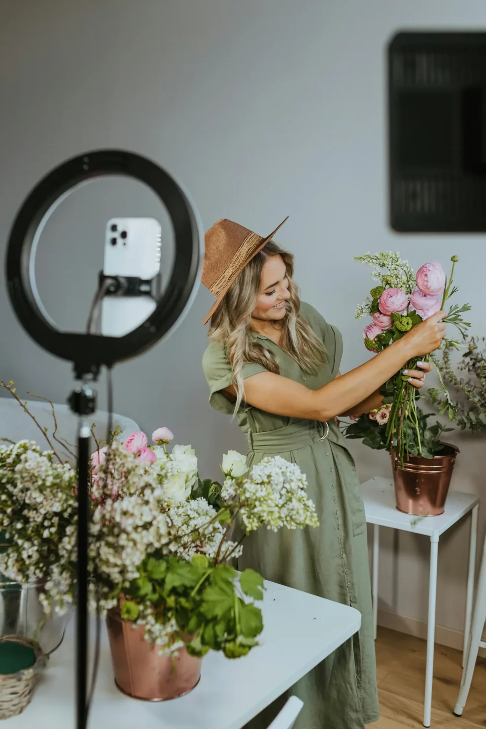Woman in tan hat arranging pink flowers while filming content with ring light and smartphone for small business social media.