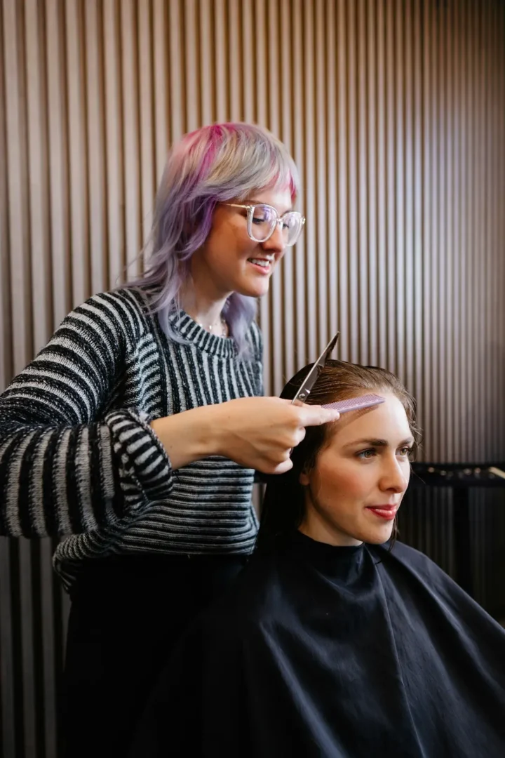 Hair stylist with pink and purple hair cutting client's hair in modern salon with textured wall backdrop.
