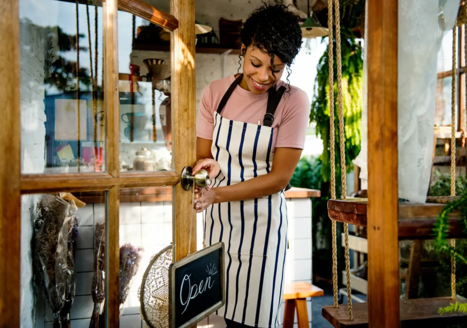 Small business owner in striped apron pouring drink at boutique shop entrance with Open sign displayed on rustic wooden door.