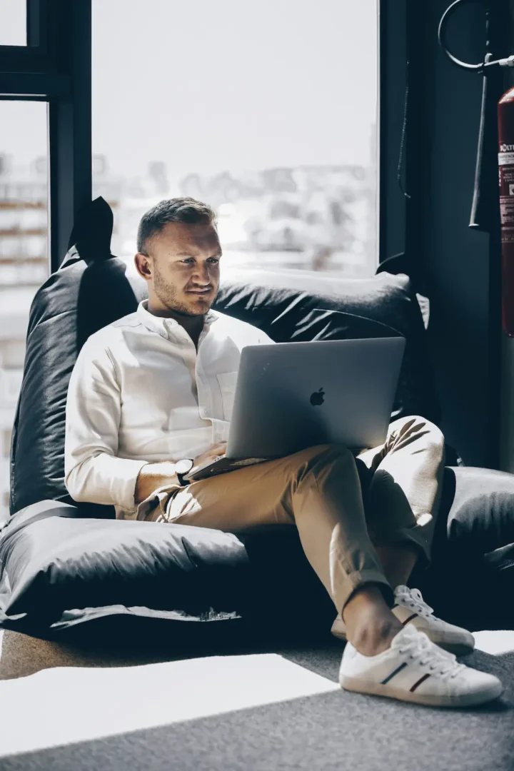 Man in white sweater working on laptop while sitting on gray couch in modern office with large windows.