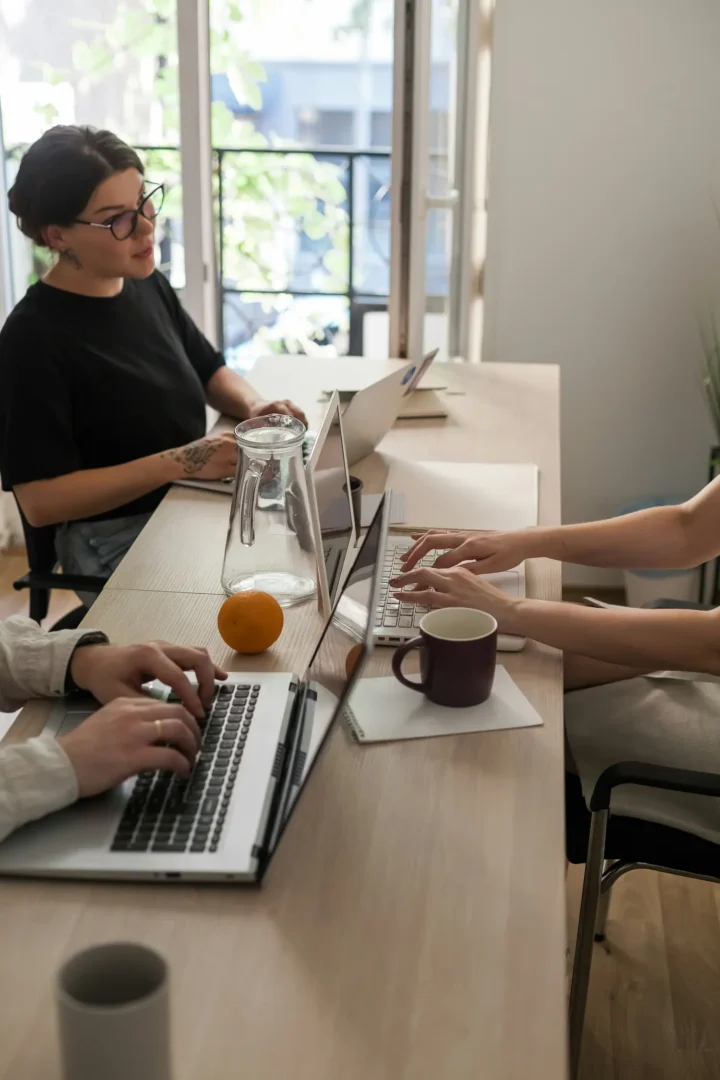 Two people working on laptops at shared wooden desk with coffee and water pitcher in bright modern workspace.
