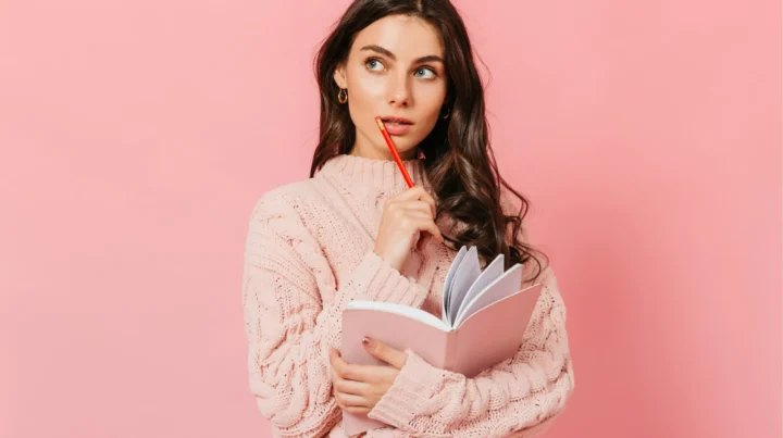 Woman in a pink sweater holding a notebook and pencil with a thoughtful expression, representing a small business owner choosing a domain name.